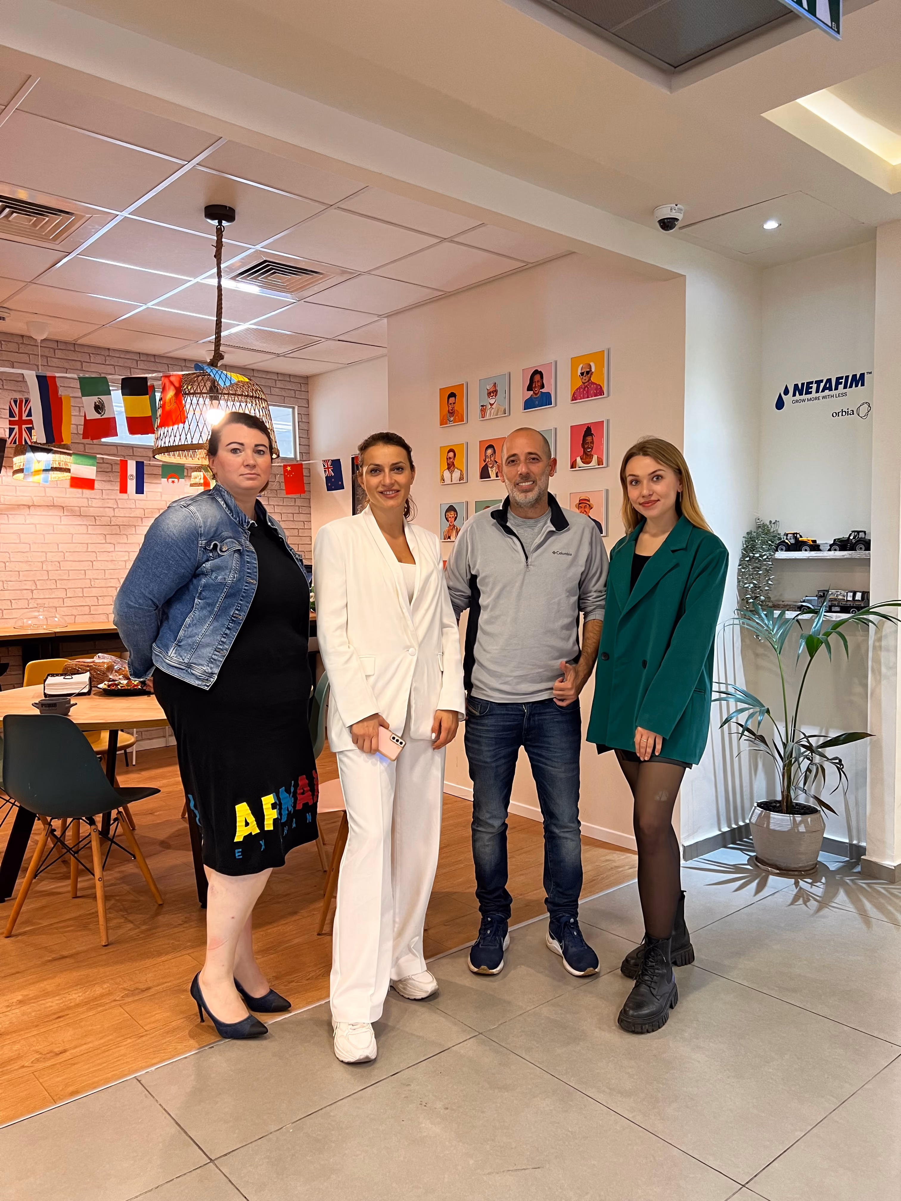Four people standing in an office with international flags, framed portraits, and a plant.