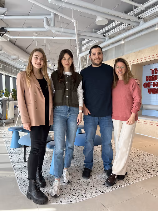 Four smiling adults standing close together in a modern office space with exposed ceilings.