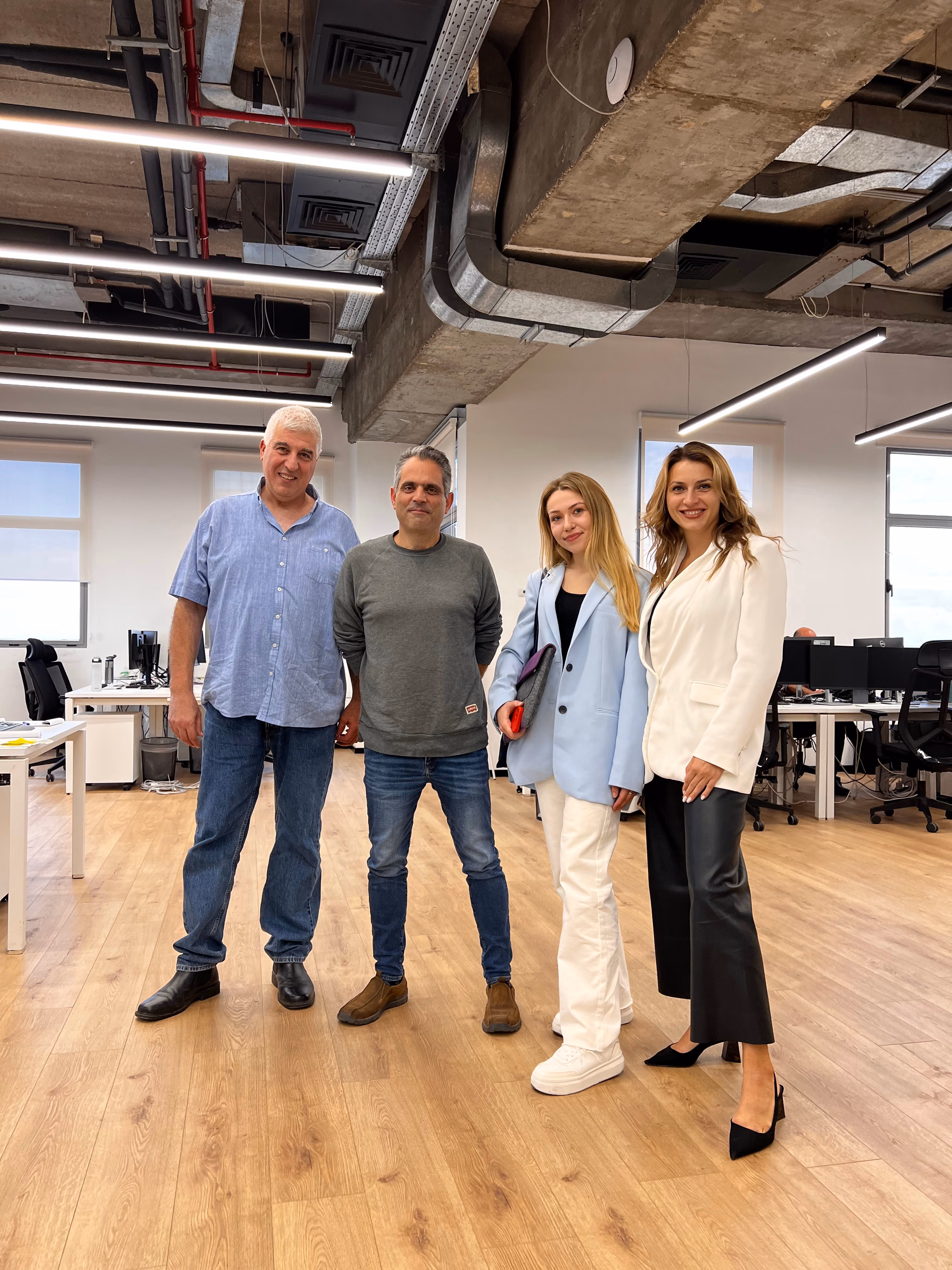 Four people standing in a modern office with wood floors and exposed ceiling pipes, smiling at the camera.