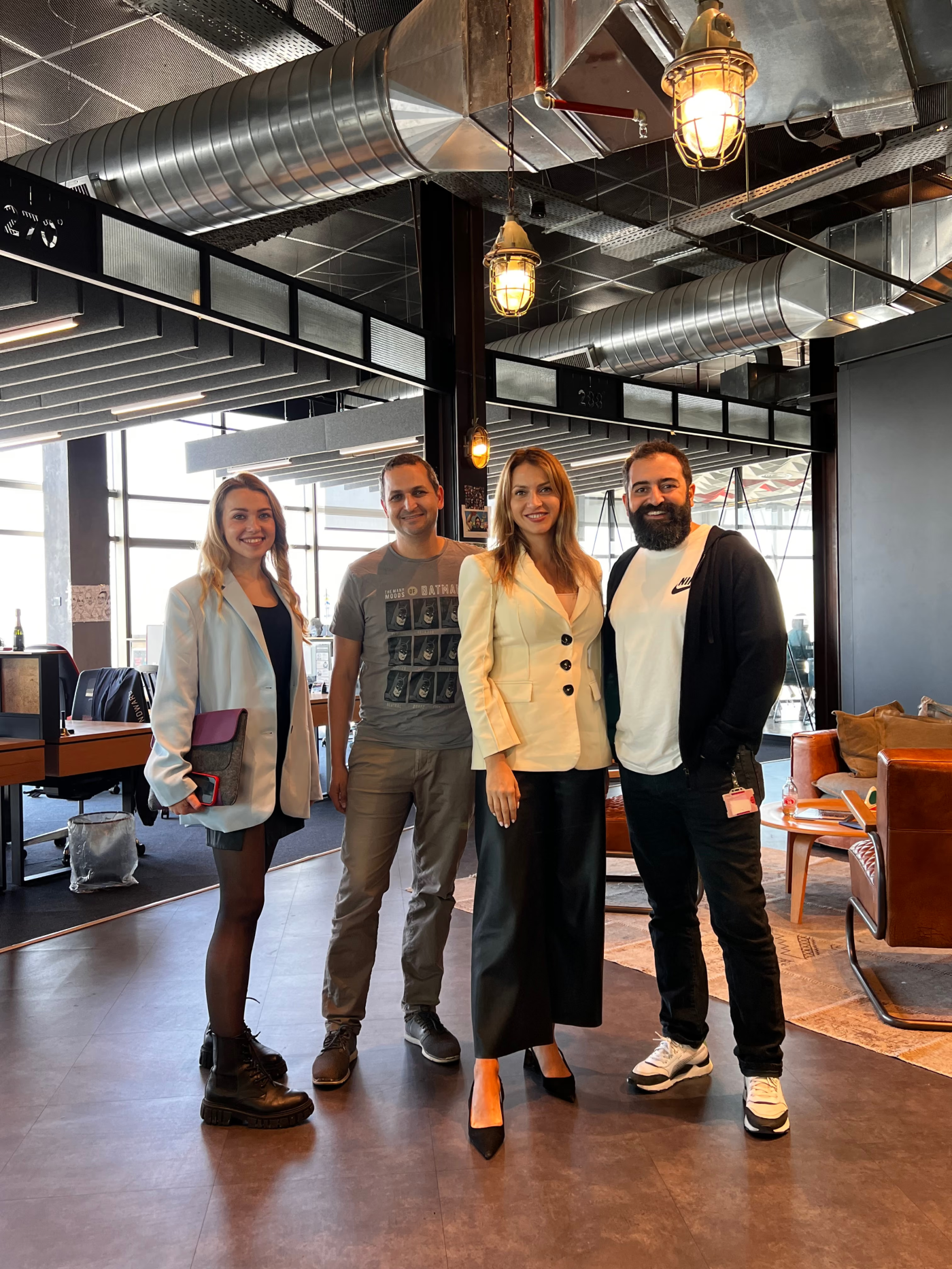 Four people standing and smiling in a modern office space with industrial ceiling design and large windows; two women wear blazers, one in a white blazer with black skirt and boots, the other in a cream blazer with black pants and heels, while two men wear casual clothes, one with a gray Batman t-shirt and gray pants, and the other with a white Nike shirt, black hoodie, black pants, and sneakers.