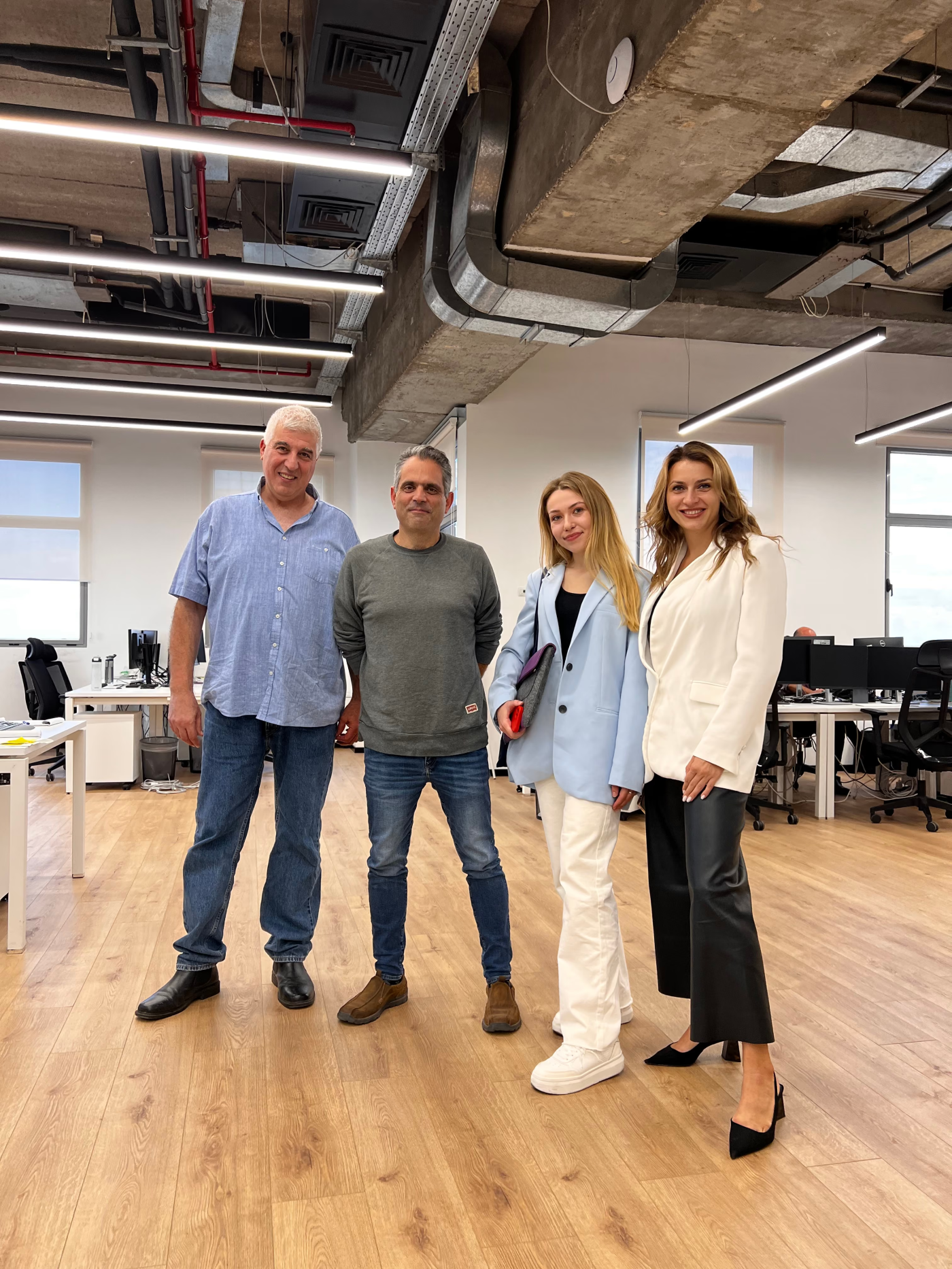 Four people standing and posing in a modern office with wooden floors and exposed concrete ceiling; two men on the left wear casual clothes—one in a light blue short-sleeve shirt and jeans, the other in a grey sweatshirt and jeans; two women on the right wear business casual, one in a light blue blazer and white pants holding a tablet, and the other in a white blazer and black pants with heels.