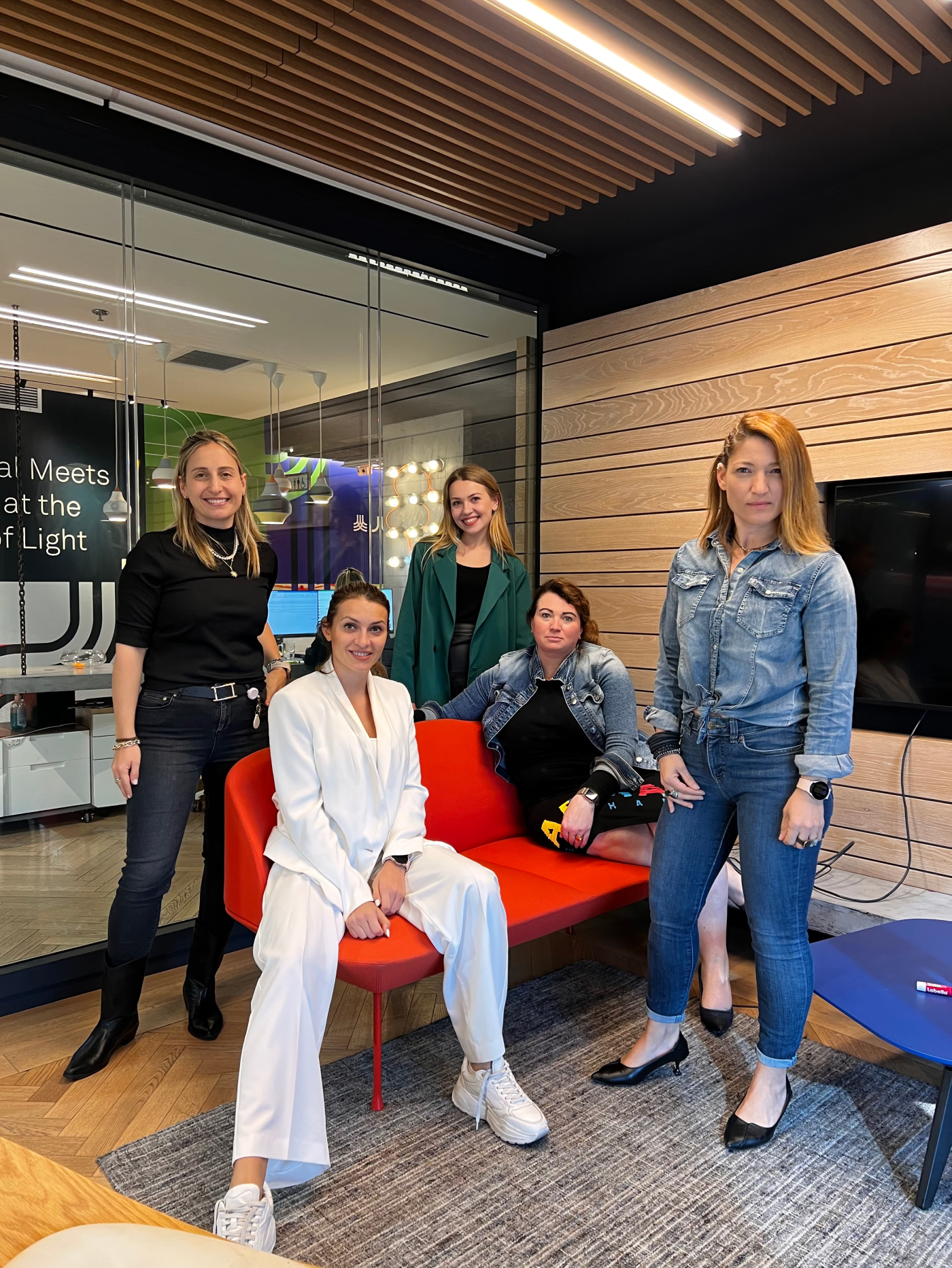 Five women posing together in a modern office with wooden wall paneling and glass partitions, three standing and two seated on a red sofa, wearing casual and business casual attire.
