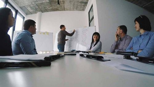 A group of people sitting around a table listening to the presenter writing on a whiteboard