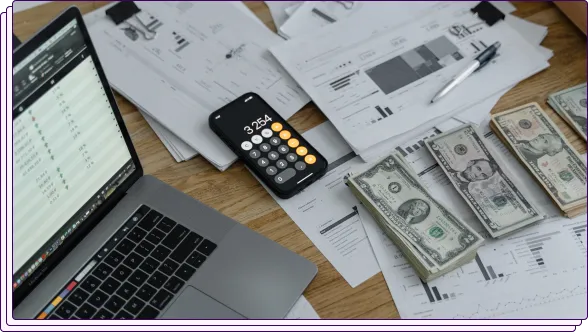 Financial paperwork, computer, calculator and piles of money on a table.