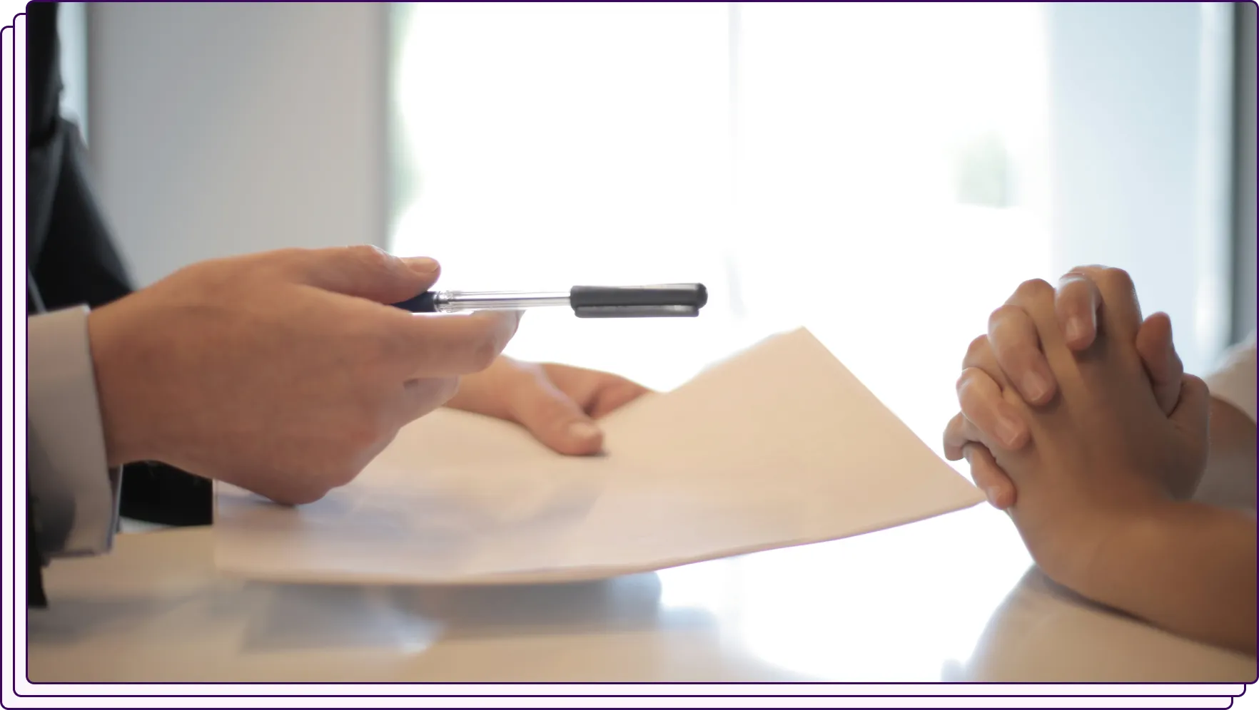 A professional handing a pen and a sheet of paper across a desk to another individual whose hands are clasped together. The image conveys an interaction involving a document review or signature, possibly in a formal setting.