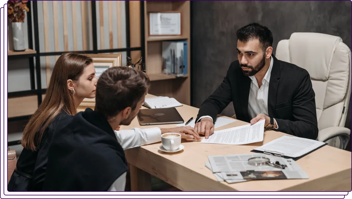 A professional in a suit sits behind a desk, holding out a document to a man and a woman seated across from him. The man appears to be pointing at the document while the woman looks on attentively. The scene suggests a discussion involving document review or legal advice in an office setting.