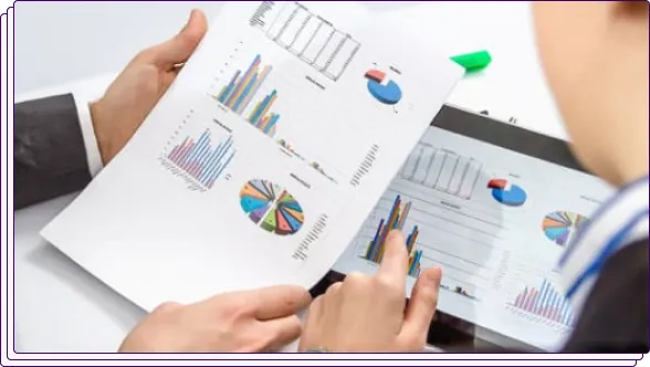 Two individuals reviewing financial charts and graphs on a paper document and a digital tablet. The visuals include colorful pie charts and bar graphs, signifying the assessment of financial data or trends.