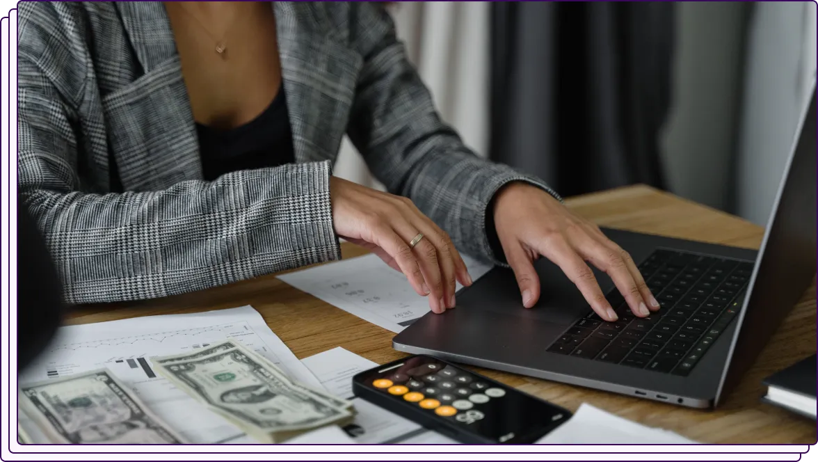 A person in a checkered blazer working on a laptop at a desk with financial documents, a calculator, and cash bills spread out. The setup highlights an environment involving financial calculations or fraud detection.
