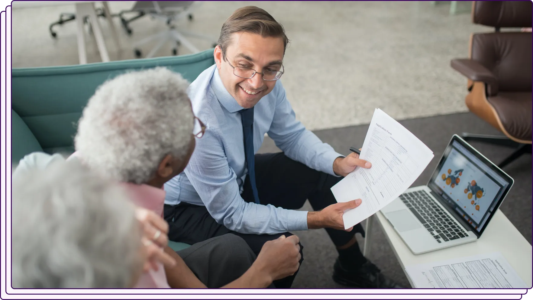 A professional with glasses and a friendly demeanor showing a document to two elderly individuals seated on a couch. A laptop displaying visual data is placed on a nearby table. The image represents a financial consultation or document review session.