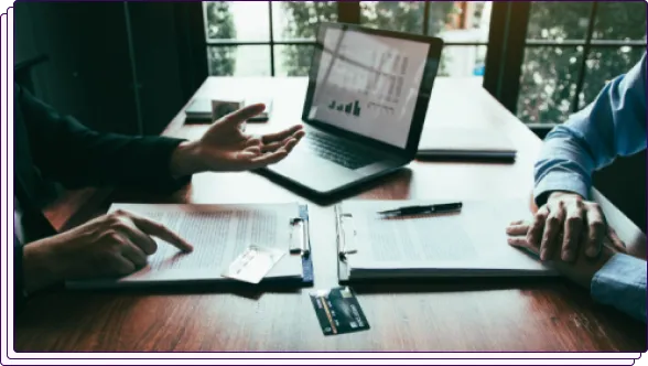 Two professionals reviewing documents on a table, with a laptop displaying charts in the background. One person is gesturing toward a clipboard with papers, while credit cards are visible on the table, signifying a financial or underwriting discussion.