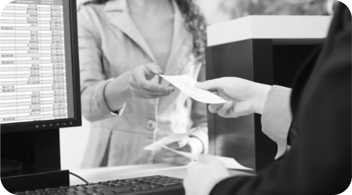 Bank teller handing a document to a customer across the counter, with payroll data on a monitor—symbolizing third-party verification and professional review of submitted pay stubs.