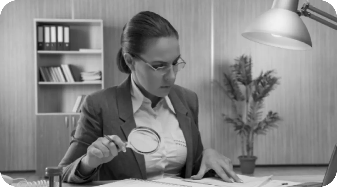 A businesswoman wearing glasses is seated at a desk under a task lamp, closely examining paperwork with a magnifying glass. Her intense focus suggests a meticulous, manual approach to document review in a professional office setting.