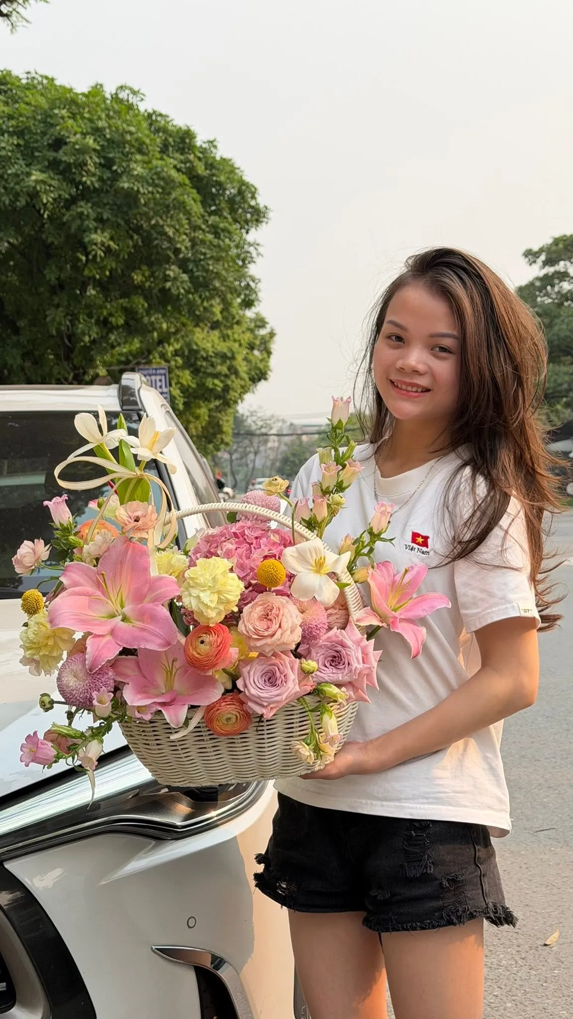 Cô gái cười cầm giỏ hoa ly hồng và hoa hồng bên cốp xe ô tô ngoài đường — King Fruit quận Tân Bình HCM