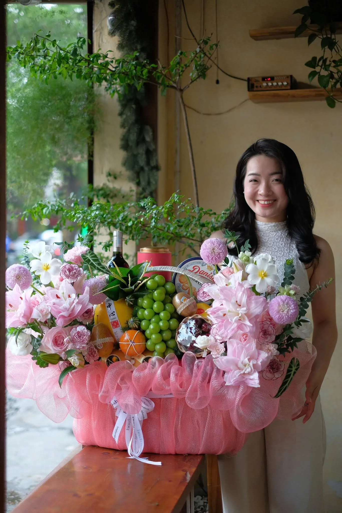 Người phụ nữ đứng cạnh giỏ hoa quả nho xanh, cam, rượu vang và hoa hồng phấn — King Fruit huyện Si Ma Cai, Lào Cai