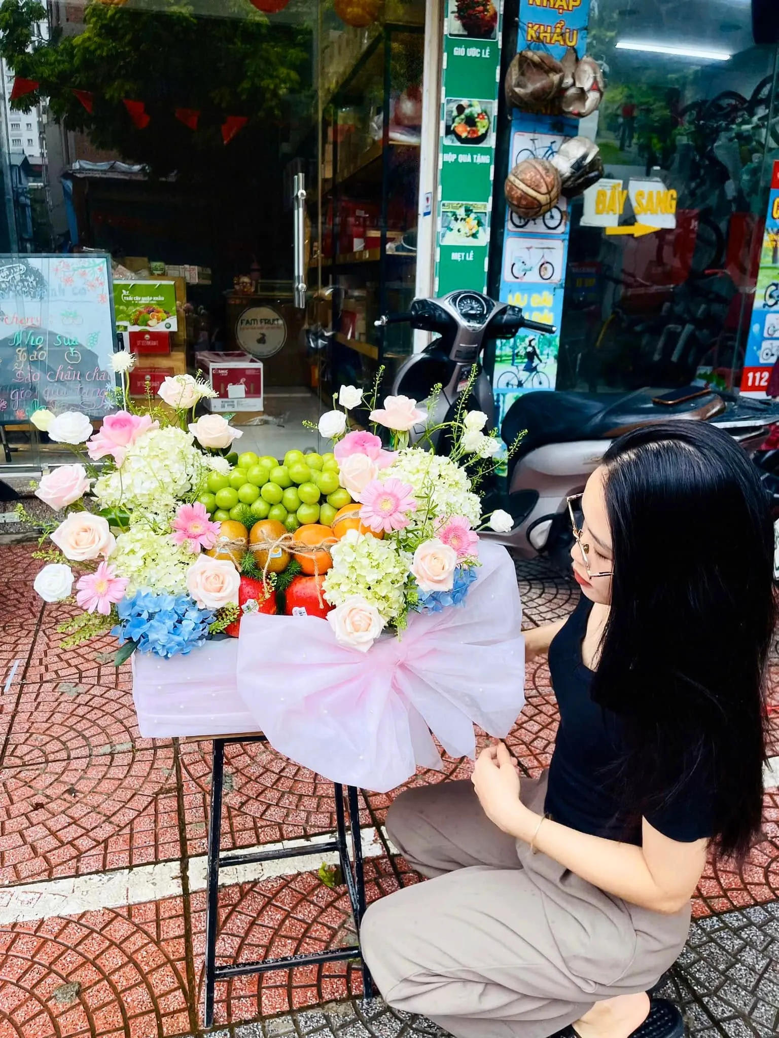 Cô gái quỳ chỉnh giỏ hoa quả nho xanh, cam, dâu tây trước cửa hàng có xe máy — King Fruit Trần Văn Thời, Cà Mau