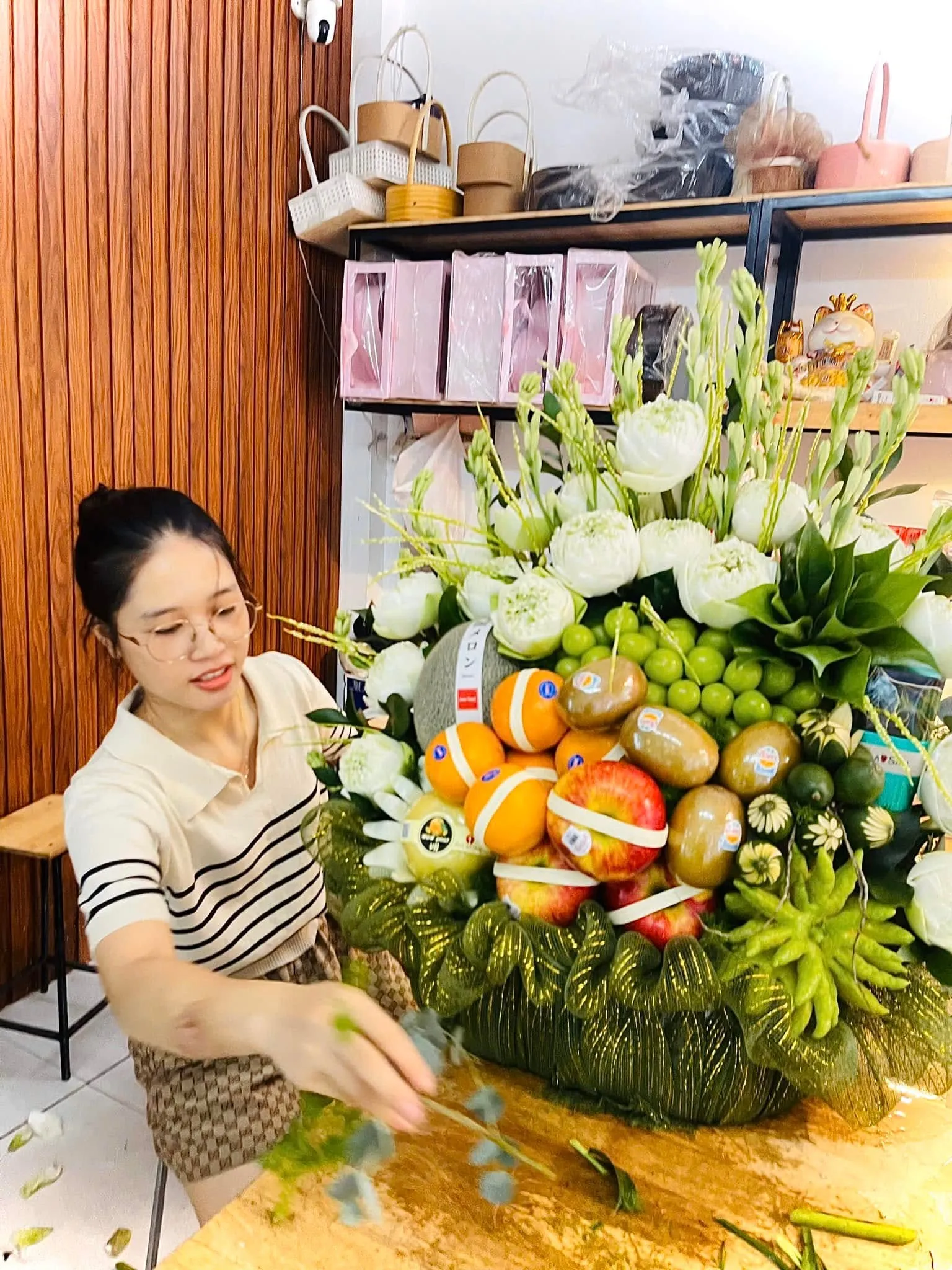 Nữ nhân viên cắm giỏ hoa quả cam, táo, nho xanh, kiwi và hoa sen trắng trên bàn gỗ — King Fruit huyện Bá Thước, Thanh Hoá