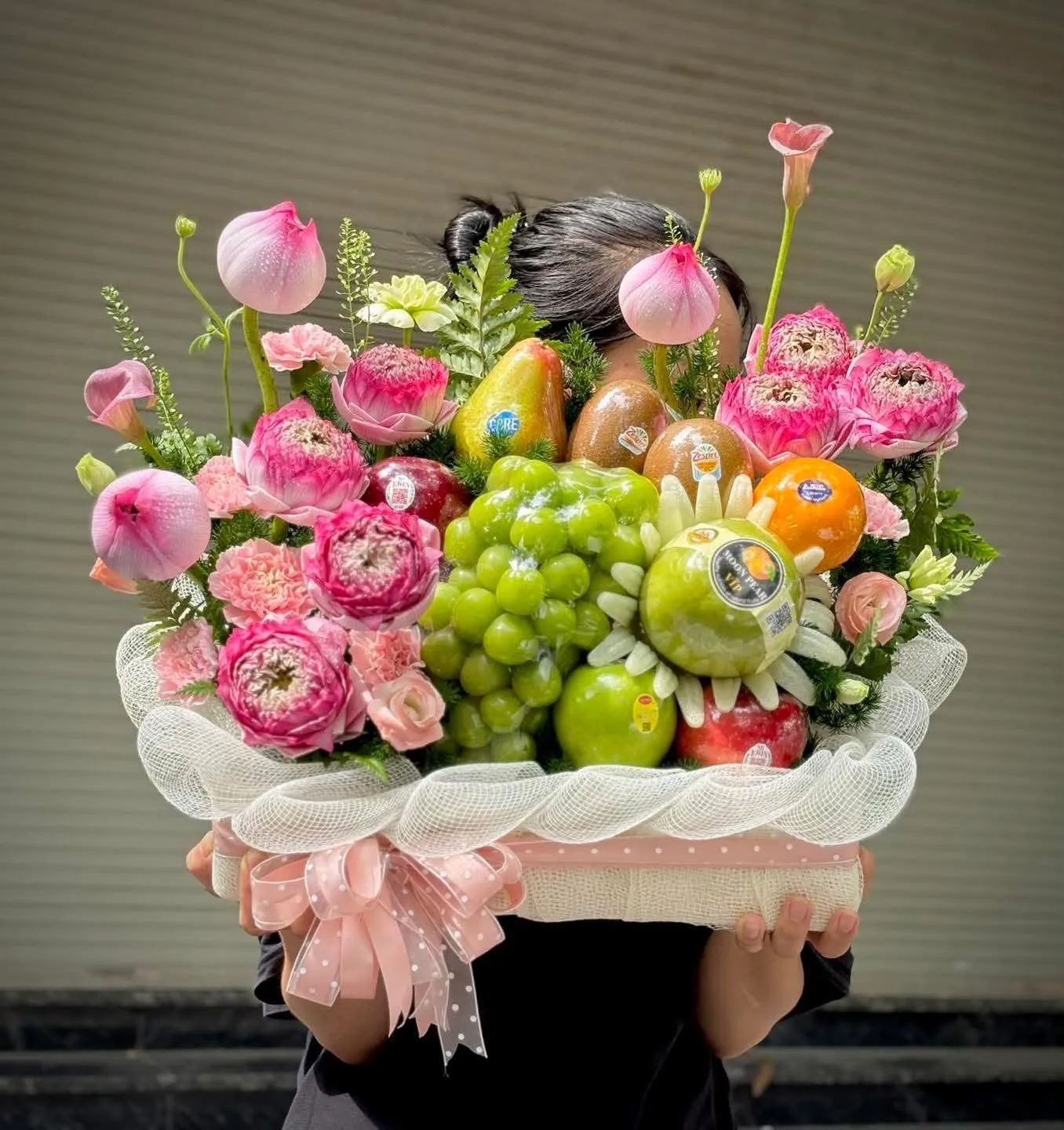 Người cầm giỏ hoa quả nho xanh, táo, lê, cam và hoa mẫu đơn hồng che khuất mặt — King Fruit Đắk Đoa, Gia Lai