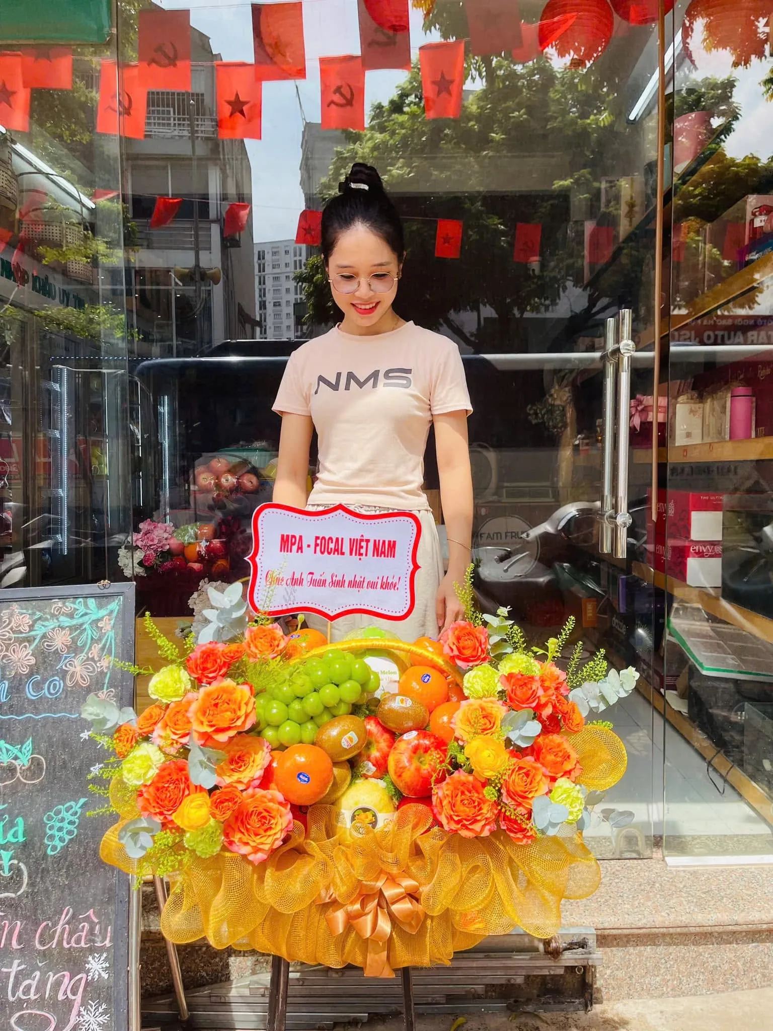 Cô gái đứng cạnh giỏ hoa quả nho xanh, cam và hoa hồng vàng trước cửa hàng — King Fruit huyện Hòn Đất, Kiên Giang