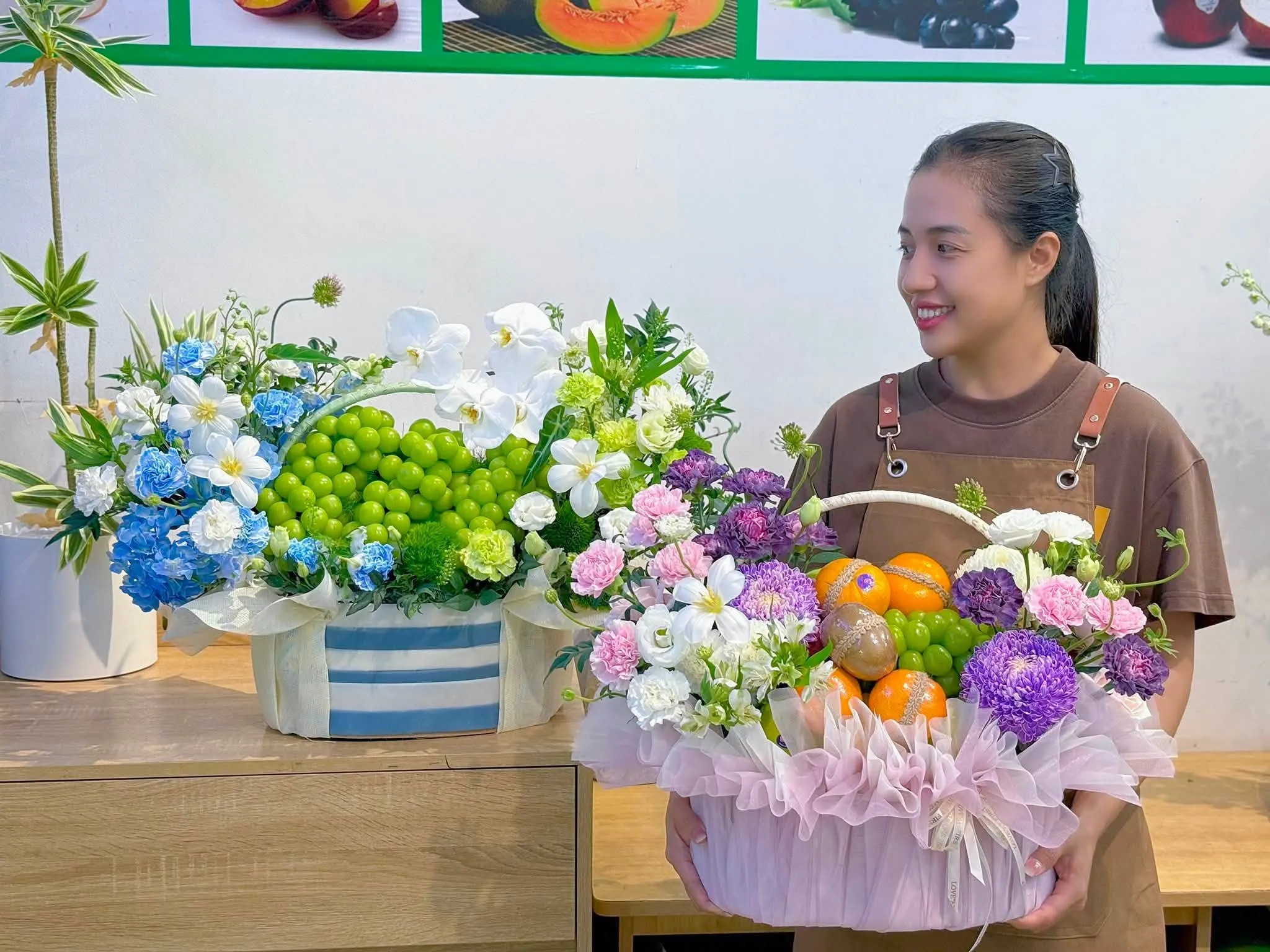 Nữ nhân viên đứng bên hai giỏ hoa quả nho xanh, cam quýt và hoa lan trắng — King Fruit thị xã Mường Lay, Điện Biên