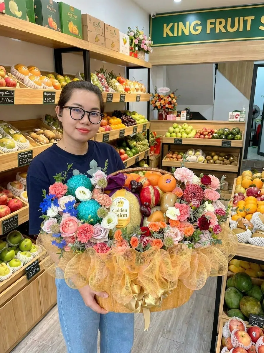 Nhân viên đeo kính cầm giỏ hoa quả có dưa lưới Golden và hoa nhiều màu tại King Fruit — King Fruit Đồng Hới - Quảng Bình