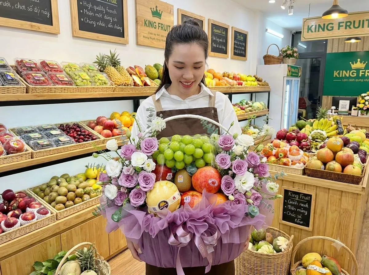 Nhân viên nữ bế giỏ hoa quả kết hoa tím giữa kệ trái cây trong cửa hàng King Fruit. — King Fruit huyện Lắk, Đắk lắk