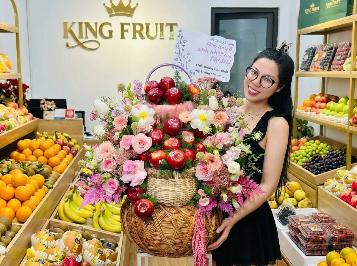Nhân viên nữ đeo kính ôm giỏ hoa quả có táo đỏ và thiệp chúc mừng trong cửa hàng King Fruit. — King Fruit huyện Lộc Bình, Lạng Sơn