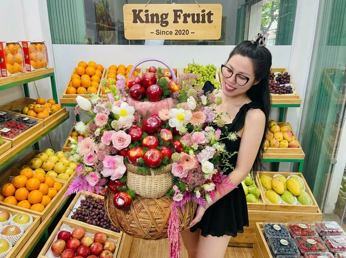 Nhân viên nữ đeo kính ôm giỏ hoa quả có táo đỏ trước kệ trái cây cửa hàng King Fruit. — King Fruit huyện Hải Hà, Quảng Ninh