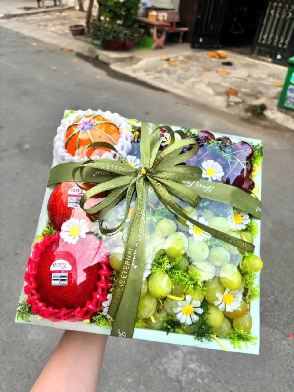 Tay cầm hộp quà trái cây có cam, xoài và nho xanh buộc nơ xanh ô liu ngoài đường phố. — King Fruit Tân Phú Đông, Tiền Giang