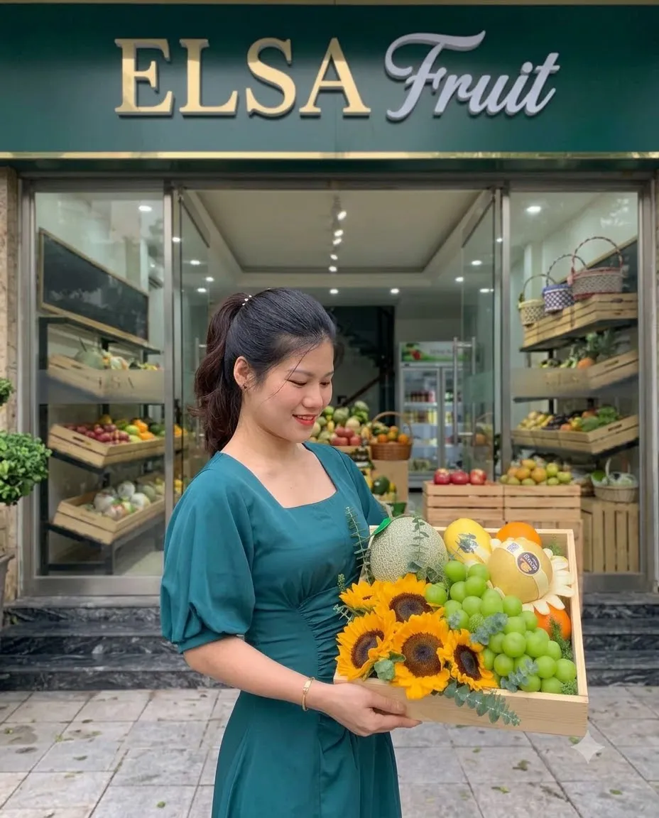 Nhân viên váy xanh cầm hộp hoa hướng dương, nho xanh và dưa lưới trước cửa ELSA Fruit — ELSA Fruit huyện Trảng Bom, Đồng Nai