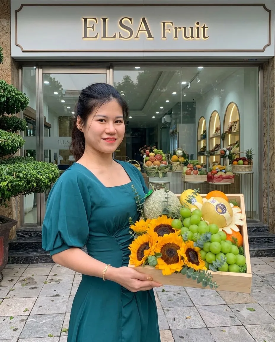Nhân viên váy xanh ôm hộp quà hoa hướng dương, nho xanh và dưa lưới trước cửa hàng ELSA Fruit — ELSA Fruit Ia Grai, Gia Lai