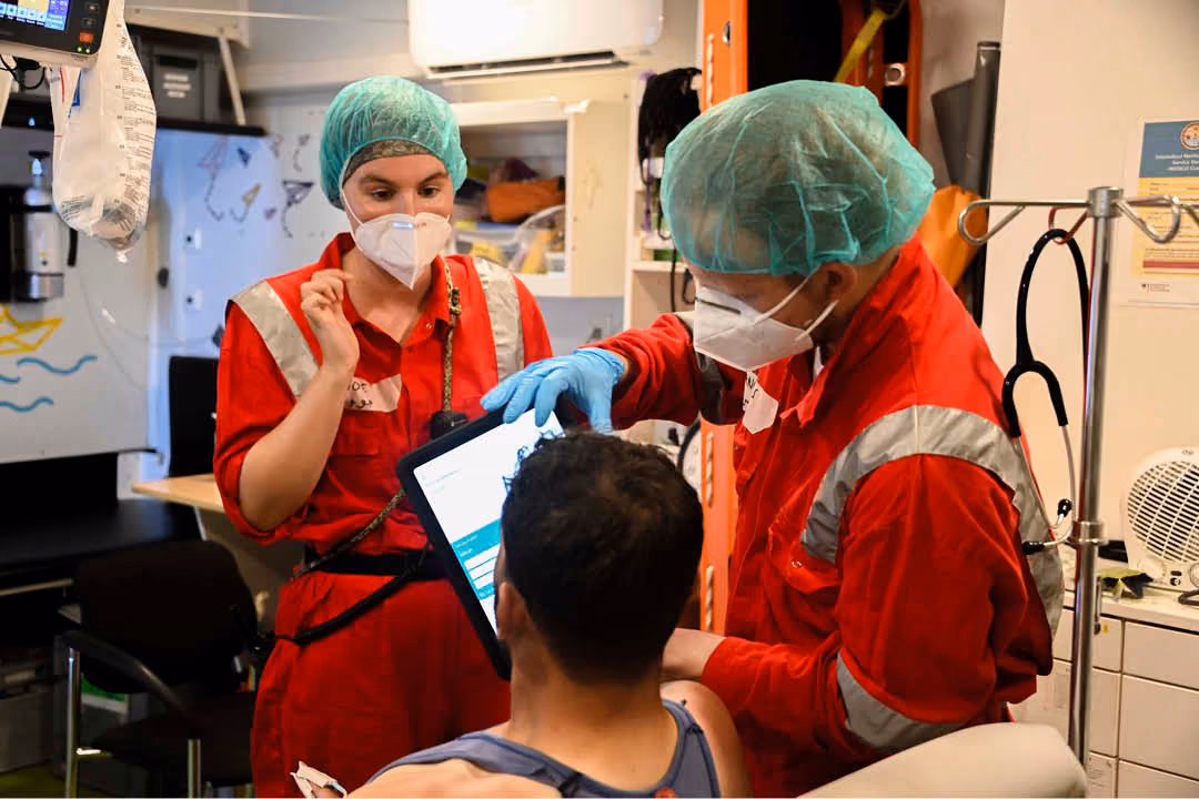 Two medical workers in red uniforms and protective gear, including face masks and hair covers, examining a patient while one holds a tablet device displaying the Care to Translate app.