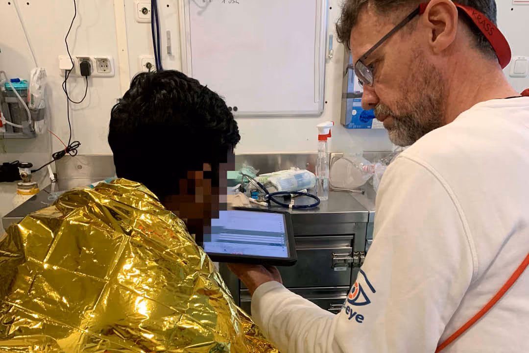 A male healthcare professional showing information on a tablet to another person wrapped in a metallic emergency blanket in a clinical setting.