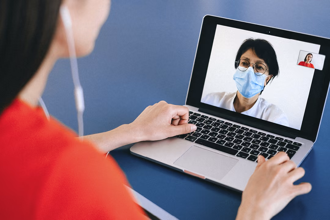 A person wearing a red shirt and earphones is having a video call on a laptop with a medical professional who is wearing glasses and a mask.