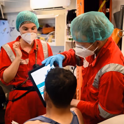 Two medical workers in red uniforms and protective gear, including face masks and hair covers, examining a patient while one holds a tablet device displaying the Care to Translate app.