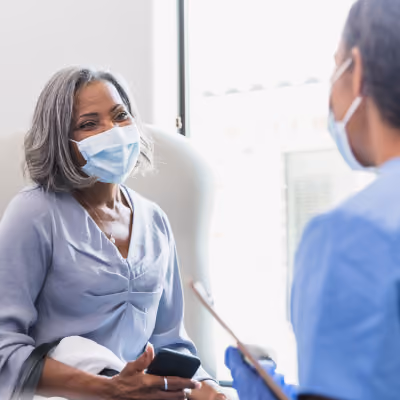Older woman wearing light purple shirt and face mask sitting in an examination room and talking to a healthcare worker in blue scrubs who is holding a clipboard.