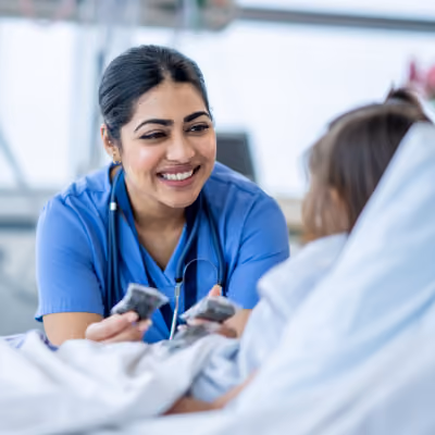 A smiling nurse in blue scrubs with a stethoscope talks to a child patient lying in a hospital bed.