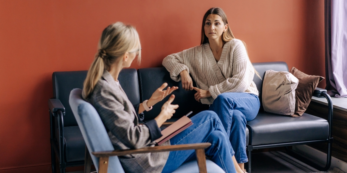 Two women with blonde hair are having a conversation on a couch. One is a therapist, one is a patient.