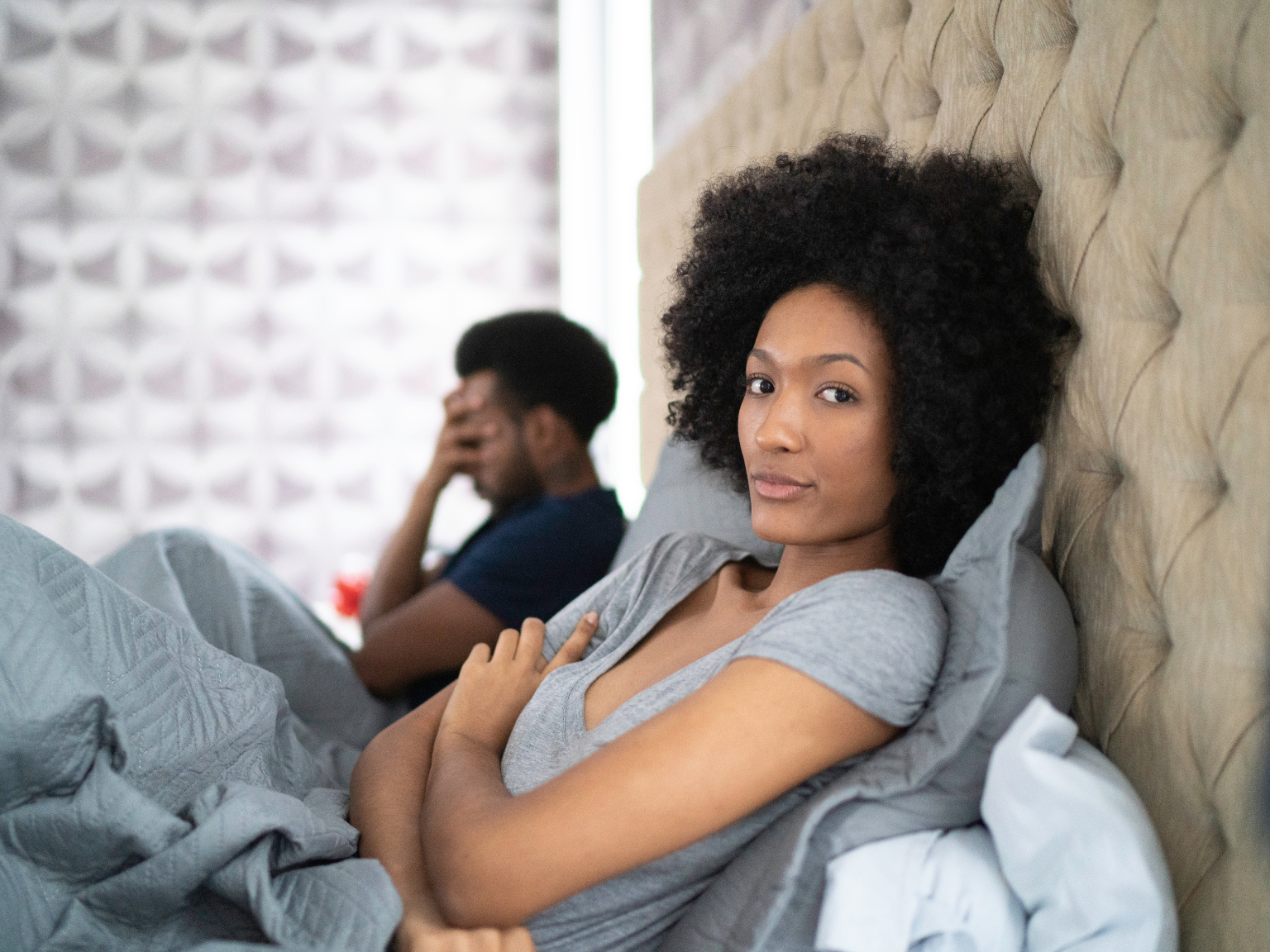 A Black couple in bed together. The man is in the background with his hand covering her face, and the woman is in the foreground, looking at the camera with a bored expression.