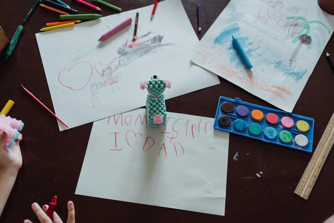 Birdseye view of a desk full of scattered coloring pencils, a watercolor palette, and three sheets of paper with children's drawings of hearts, palm trees, the beach and the ocean, as well as some early writing practice of the words mom and dad.