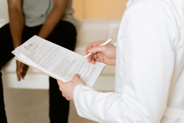 A person sitting in a doctor's office while the doctor reviews medical history on a clipboard.