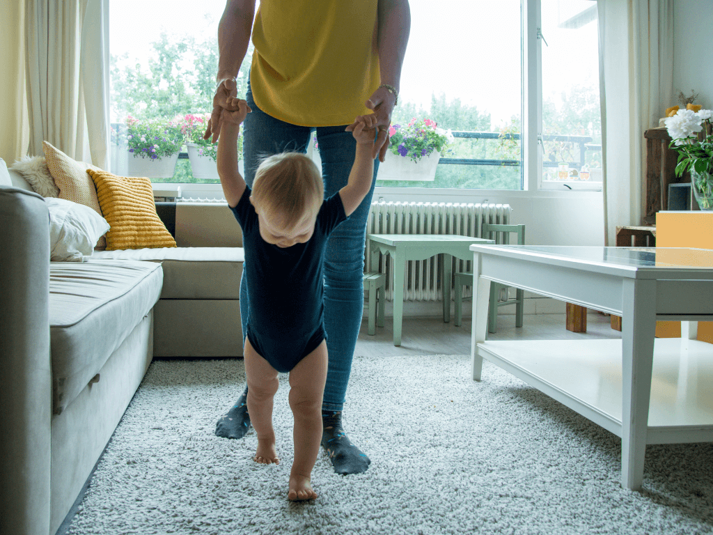 An adult and toddler in a living room; the adult is holding onto the toddler's hands and keeping them steady while they practice walking.