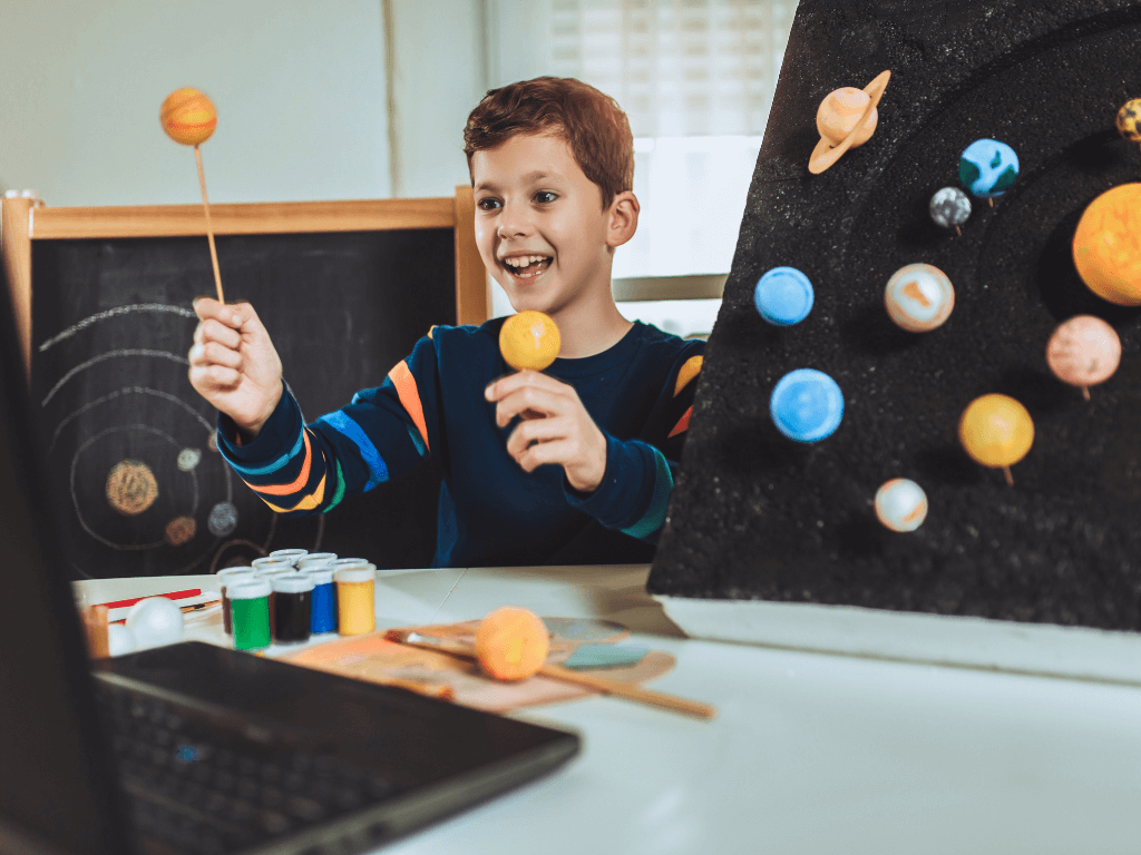 An autistic child excitedly holding colorful planet models while they work on homework. The table they're sitting at has paint, more models, and a laptop.