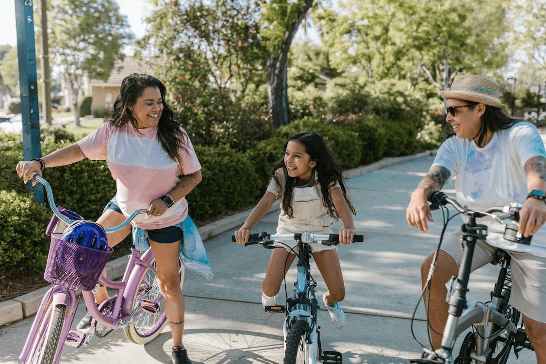 A BIPOC rainbow family of three smiling at each other while riding their bikes in a park.