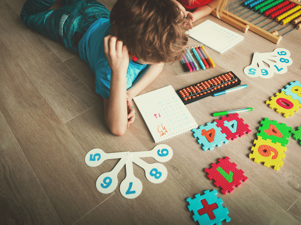 A young boy lying on the floor looking at various colorful number learning puzzles and visual aids.