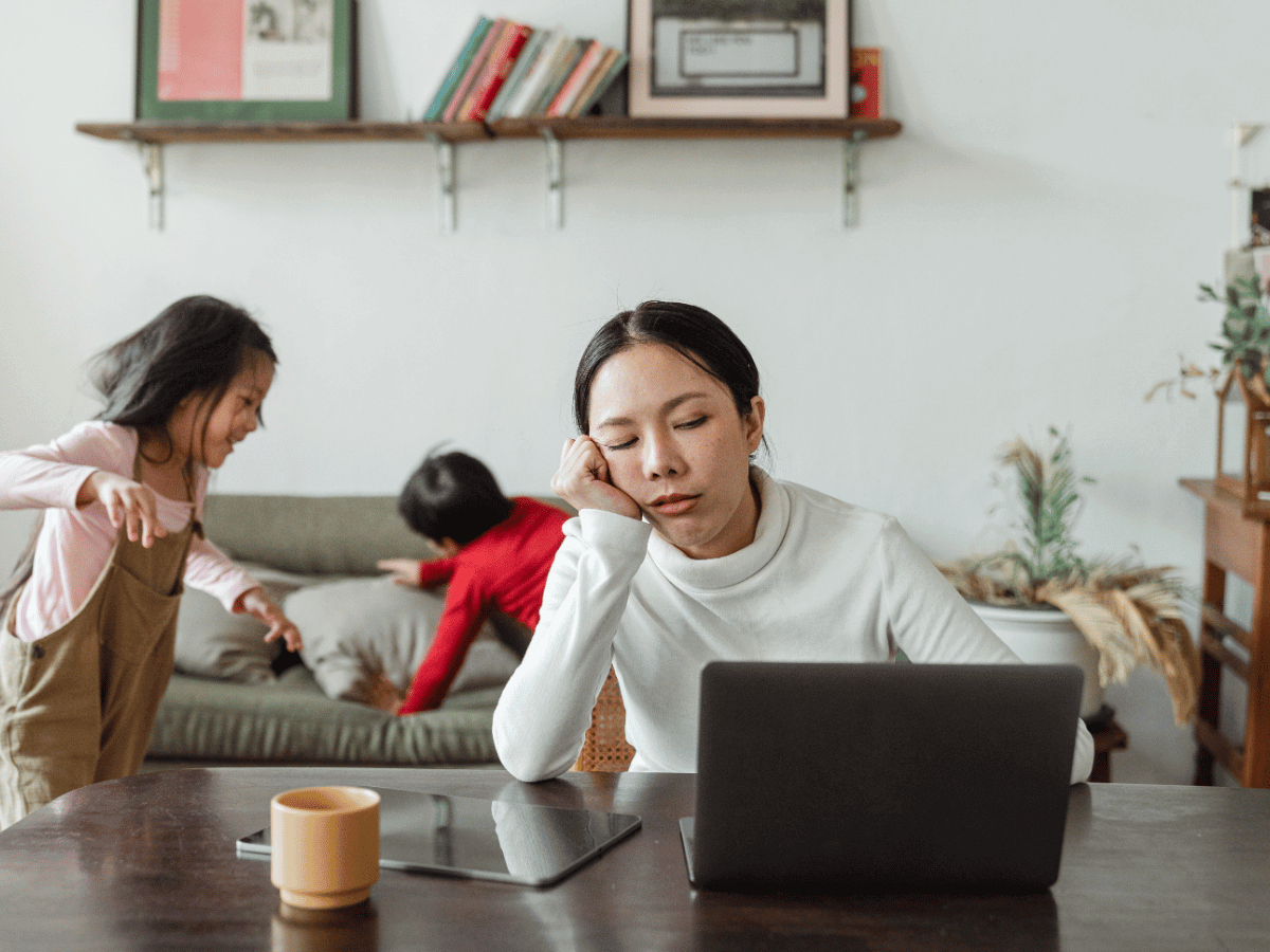 A woman with ADHD is exhausted and tired. She has kids playing in the background while she works at home with coffee.