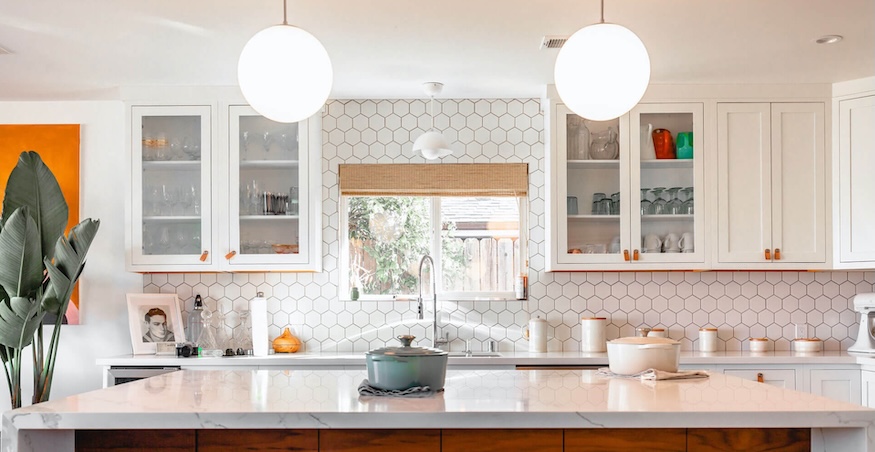 Bright modern kitchen with white hex tile backsplash, marble island, hanging round lights, and plants on left.