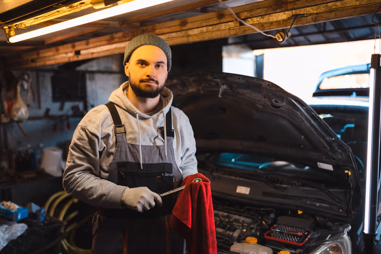 NH Garage Keepers Insurance concept - Waist-up portrait of a young mechanic holding a socket wrench in front of a car with open hood in his garage