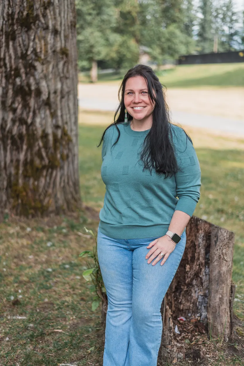 Krystal standing outdoors near a tree, smiling while wearing a teal sweater and jeans.