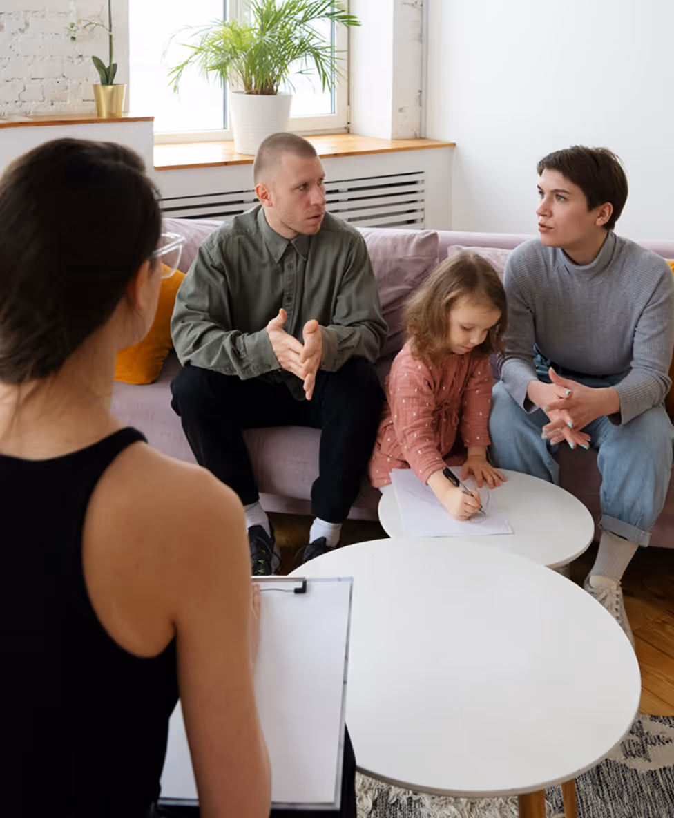 A family therapy session with two adults talking, a child writing, and a therapist observing.