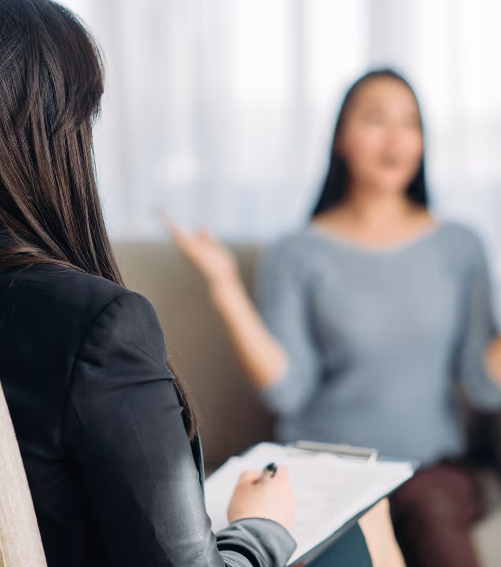 A therapy session with a woman speaking while another takes notes on a clipboard.
